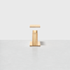A small, modern wooden pedestal stands on a light gray floor against a plain white background. The pedestal has clean lines and a minimalist design.