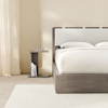 A modern, minimalist wooden nightstand with a flat top and base, positioned beside a bed with white bedding against a white wall. The nightstand has a simple C-shaped design and a smooth, light brown finish.