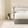 A modern, minimalist wooden nightstand with a flat top and base, positioned beside a bed with white bedding against a white wall. The nightstand has a simple C-shaped design and a smooth, light brown finish.