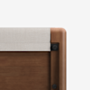 Close-up of the corner of a wooden bed frame with rounded edges and a light beige fabric headboard panel against a white background.