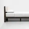 Side view of a modern bed with a dark wooden frame, a beige upholstered headboard, and white bedding, set against a plain white wall and floor.