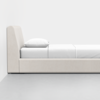 A modern, minimalist bed with a light beige upholstered frame, a white mattress, white bedding, and a single white pillow, positioned against a plain white wall on a gray floor.