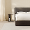 A modern, dark wood nightstand with a geometric design sits next to a wooden bed frame with white bedding in a minimal, white-walled room.