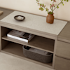 A light brown wooden sideboard with a beige stone top holds a brown vase with flowering branches, a dark plate with decorative objects, books on the shelf below, and a gray felt basket on the bottom shelf.