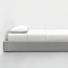 A minimalist gray platform bed with a white mattress, white sheets, and a single white pillow, set against a plain white wall and floor.