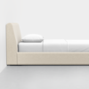 A minimalist beige upholstered bed with a tall headboard, white bedding, and a single white pillow, set against a plain white wall and gray floor.