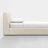 A minimalist beige upholstered bed with a tall headboard, white bedding, and a single white pillow, set against a plain white wall and gray floor.