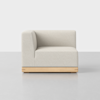 A modern, minimalist corner sofa chair with light beige upholstery and a wooden base, set against a plain white background on a light grey floor.