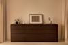 A dark wooden dresser against a neutral wall, topped with two small potted plants, a framed abstract artwork, and a white candle. Light curtains frame the minimalist, softly-lit scene.