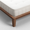A close-up view of the corner of a bed with a wooden frame and a white, tufted mattress on top, placed on a light-colored floor.