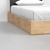 Close-up of a wooden bed frame with a light natural finish, white bedding, and a white pillow, positioned on a smooth, light gray floor against a white wall.