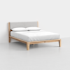 A modern bed with a light wooden frame, angled legs, a light gray upholstered headboard, and white bedding with two pillows, set against a plain white wall and floor.