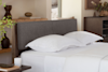 A modern bedroom with a gray upholstered headboard, white pillows, and white bedding. Wooden shelves and decor items such as vases and books are visible in the background, creating a calm, minimalist atmosphere.