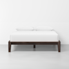 A minimalist bed with a dark wooden frame, white bedding, and two white pillows, set against a plain white wall and light gray floor.