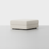 A minimalist, square, cream-colored ottoman with a soft, textured fabric and low black feet, placed against a plain light gray background.