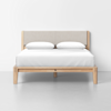 A minimalist wooden bed frame with a padded beige headboard, made up with white sheets and two white pillows, is set against a plain white wall and gray floor.