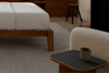 A cozy modern bedroom with a wooden bed frame, white bedding, a textured armchair, a small wooden side table holding a ceramic mug, and a wooden stool in the background on a large rug.
