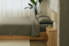 A minimalist bedroom with a wooden bed frame, gray bedding, light wood nightstand, beige lamp, potted plant by the window, and sheer white curtains letting in natural light.