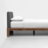 A minimalist bed with a dark upholstered headboard, white bedding, and a wooden frame positioned against a white wall on a light-colored floor.