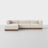 A modern, light beige sectional sofa with a left-facing chaise and low wooden base, placed on a light gray floor against a plain white background.