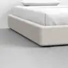 A close-up view of a modern bed with a beige upholstered frame and white bedding, set on a smooth, light gray floor against a plain white wall.