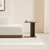 A modern wooden bed with white bedding is positioned next to a small, matching wooden nightstand against a plain white wall and light gray floor.