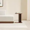 A modern wooden bed with white bedding is positioned next to a small, matching wooden nightstand against a plain white wall and light gray floor.