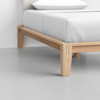 A close-up view of a wooden bed frame with light wood legs and a white mattress, set on a smooth, light gray floor next to a white wall.