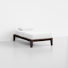 A minimalist single bed with a dark wooden frame, white mattress, white bedsheet, and a white pillow, positioned in an empty, light-colored room.