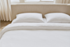 A neatly made bed with two white pillows and a white textured comforter, set against a beige upholstered headboard. Sunlight streams in through windows with light-colored curtains in the background.