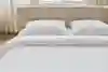 A neatly made bed with two white pillows and a white textured comforter, set against a beige upholstered headboard. Sunlight streams in through windows with light-colored curtains in the background.
