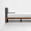 A minimalist bed with a wooden frame, dark upholstered headboard, and white bedding stands against a plain white wall and floor in a modern, uncluttered room.