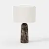 A table lamp with a cylindrical white fabric shade and a dark, marble-patterned base featuring a round metallic button near the bottom. The lamp is set against a plain white background.