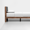 A minimalist wooden bed with a gray upholstered headboard, white bedding, and a single white pillow, positioned against a plain white wall and floor.