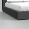 A close-up view of the corner of a bed with a dark fabric frame and white bedding, set on a light grey floor.