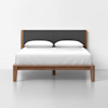 A wooden bed frame with a dark gray upholstered headboard, two white pillows, and a neatly made white bedspread, set against a plain white wall and light gray floor.