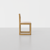 A minimalist wooden chair with a square, geometric design, shown in side profile against a plain white background and light grey floor.