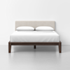 A minimalist bed with a beige upholstered headboard, two white pillows, and white bedding on a dark wooden frame, set against a plain white wall and floor.