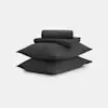 Two large dark gray pillows are stacked on top of each other with neatly folded matching dark gray bed sheets placed on top, all set against a plain white background.