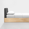 A minimalist bed with a light wood frame, white bedding, and a dark gray upholstered headboard, set against a plain white wall and gray floor.