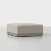 A square, light beige upholstered ottoman with a wooden base, placed on a smooth, light-colored surface against a plain white background.