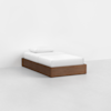 A minimalist single bed with a wooden frame, white mattress, white pillow, and white bedding placed in a bright, empty room with light-colored floors and walls.