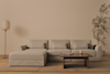 A minimalist living room with a light gray sectional sofa, a wooden coffee table with a book and mug, a floor lamp, a beige rug, and a tall potted plant against a neutral wall.