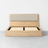 A minimalist wooden bed frame with a light wood finish, a wide headboard featuring a beige upholstered cushion, and wooden slats, shown without a mattress against a plain white wall.