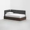 A modern daybed with a dark wooden frame, white mattress, and gray upholstered backrest, positioned in the corner of a minimalist white room.
