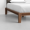 Close-up of the corner of a wooden bed frame with white bedding and a white pillow, set on a light gray floor against a plain white wall.