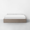 A modern bed with a light wood frame, white sheets, and two white pillows sits centered against a plain white wall on a light gray floor.