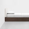 A minimalist side view of a bed with a wooden base, white bedding, a single white pillow, and an upholstered white headboard against a plain white wall.
