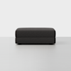 A modern, rectangular, dark gray ottoman with a simple, minimalist design is placed on a light gray floor against a plain white background.