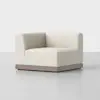 A modern, cream-colored corner armchair with a minimalist design and a wooden base, placed on a smooth, light gray floor against a plain white background.